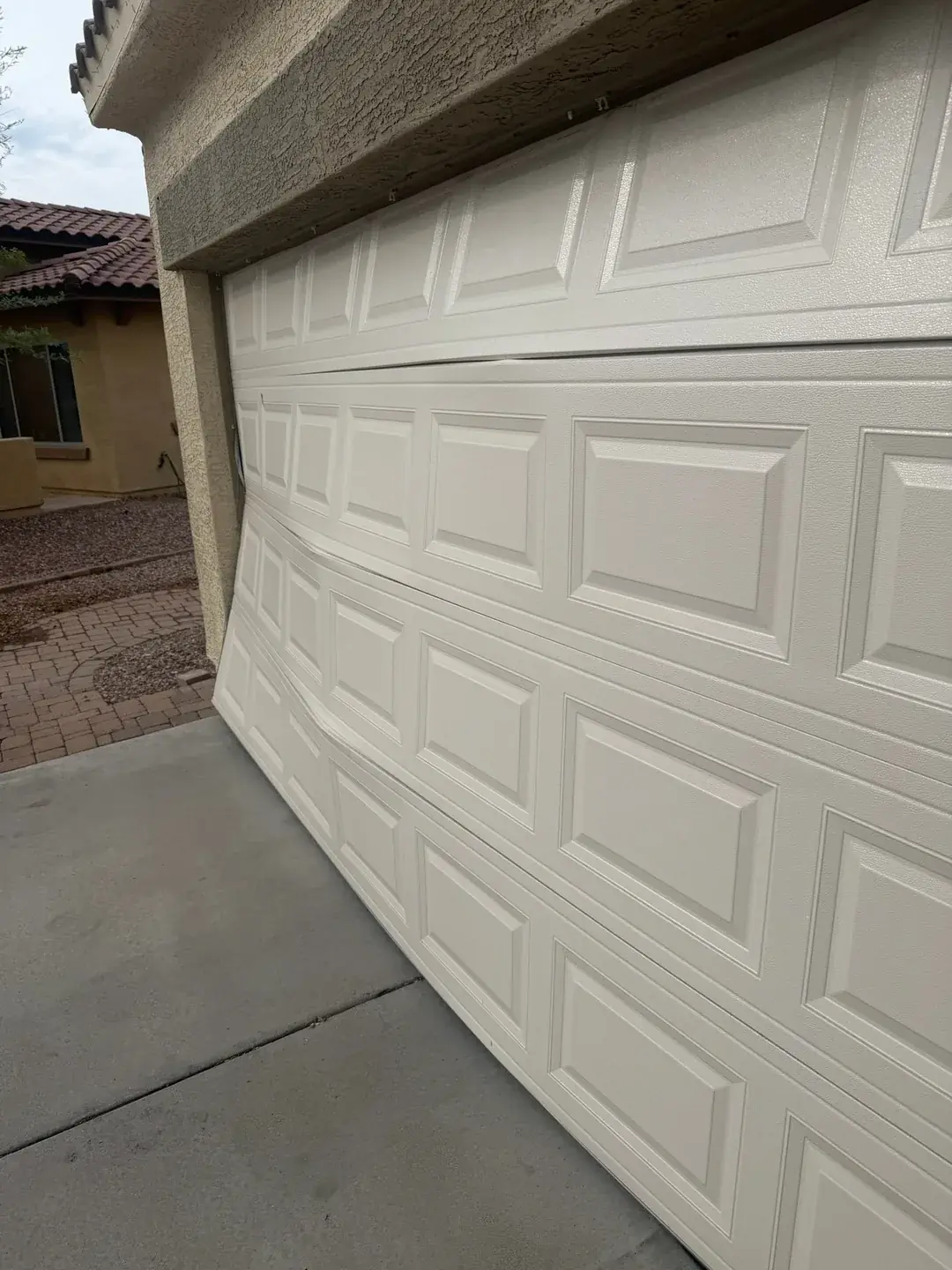 A closed cream raised-panel garage door seen at an angle from a paver driveway, bowed outward so that its top section stands clear of the header and a gap runs down the jamb, on a stucco house with a clay tile roof.